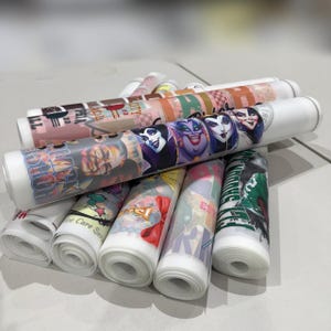 May include: A collection of rolled heat transfer vinyl sheets featuring various colorful graphic designs, including illustrations of animated characters, text, and patterns. The translucent rolls are stacked together, displaying vibrant prints.