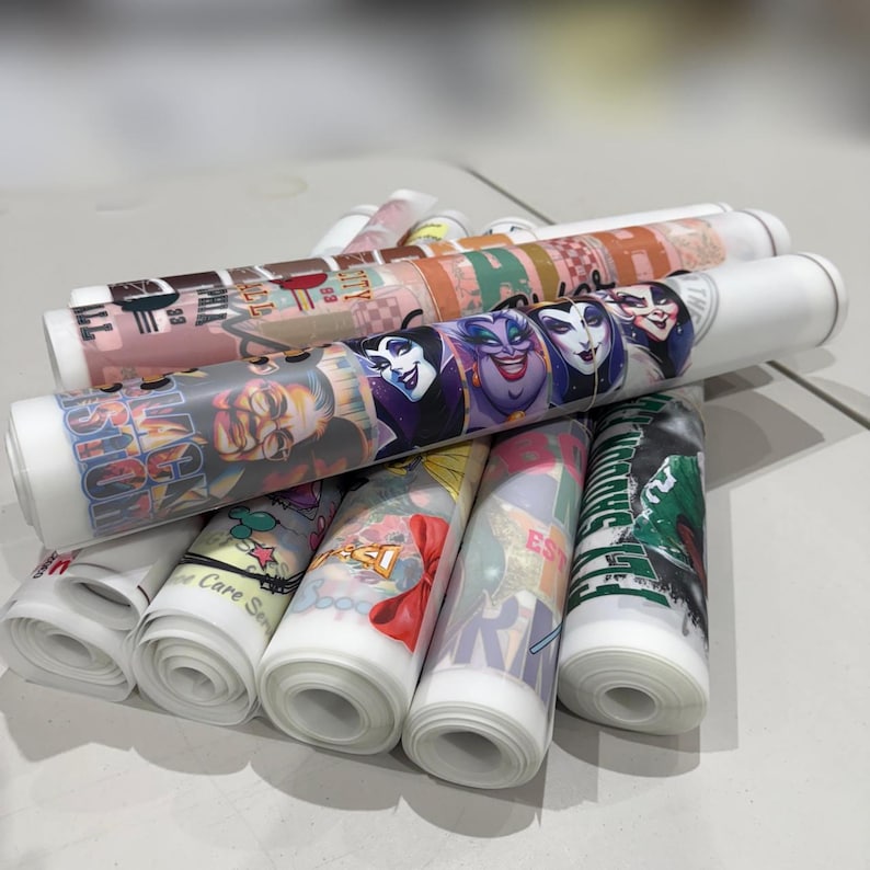 May include: A collection of rolled heat transfer vinyl sheets featuring various colorful graphic designs, including illustrations of animated characters, text, and patterns. The translucent rolls are stacked together, displaying vibrant prints.