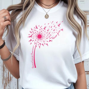 May include: A white t-shirt featuring a pink graphic of a dandelion where the seeds are small breast cancer awareness ribbons. The text "Breast Cancer Awareness" runs along the stem, and the words "Never give up" are printed in pink script.