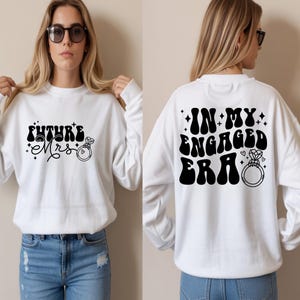 May include: White sweatshirt featuring the text "FUTURE Mrs" on the front and "IN MY ENGAGED ERA" on the back in black. The design includes a diamond ring graphic and star accents.