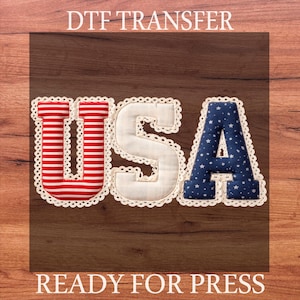 Peut inclure: Transfert DTF avec le mot "USA" en rayures rouges et blanches, blanc et bleu avec des étoiles blanches. Les lettres sont bordées d'une bordure en dentelle blanche. Les mots "DTF TRANSFER" et "READY FOR PRESS" sont imprimés.