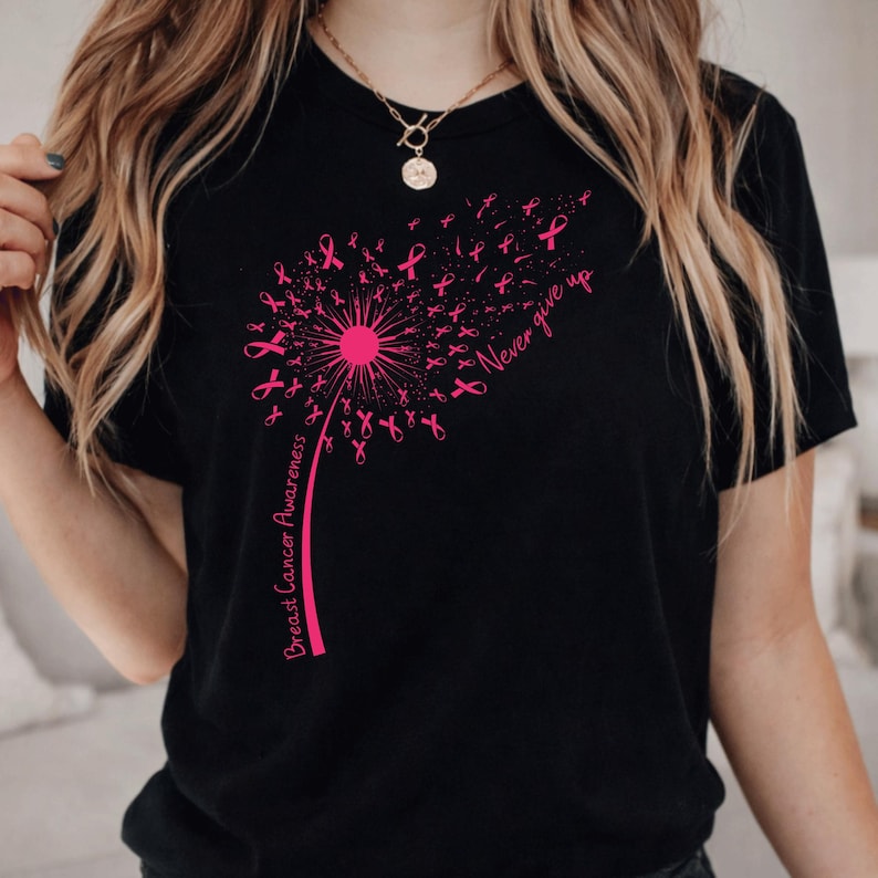 May include: A black short-sleeved t-shirt featuring a pink graphic of a dandelion where the seeds are small breast cancer awareness ribbons. The text "Breast Cancer Awareness" and "Never give up" are printed in pink script on the shirt.