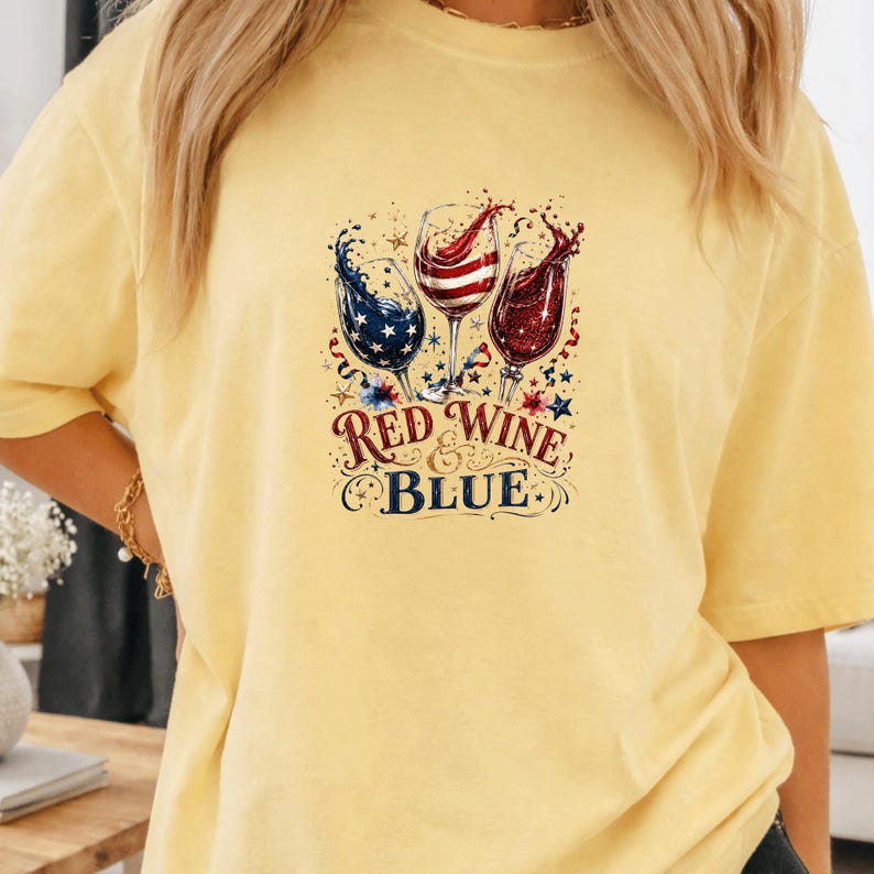 May include: A pale yellow t-shirt featuring a graphic of three wine glasses filled with red, white, and blue liquid, decorated with stars and swirls. The text "RED WINE & BLUE" is printed in a decorative font below the wine glasses.