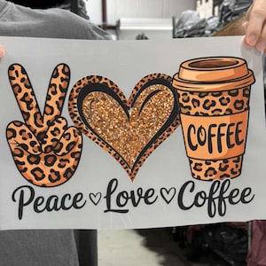 May include: A white rectangular transfer sheet featuring a leopard print peace sign, a glittery gold heart, and a leopard print coffee cup. Below the graphics, the black text reads Peace, Love, and Coffee, separated by small black heart icons.