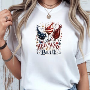 May include: A white t-shirt featuring a graphic of three wine glasses filled with red, white, and blue liquid, accented with stars and swirls. Below the glasses, the text reads RED WINE & BLUE in a decorative font, surrounded by small star motifs.