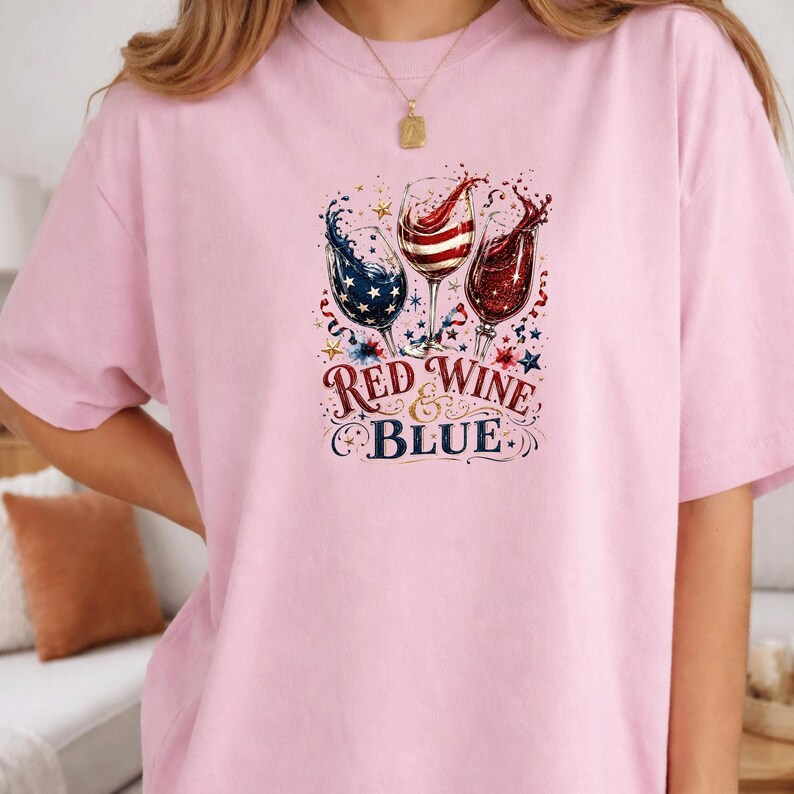 May include: A light pink t-shirt featuring a graphic of three wine glasses filled with red, white, and blue liquid. The design includes star patterns and the text "RED WINE & BLUE" in a decorative font, surrounded by small stars and confetti.