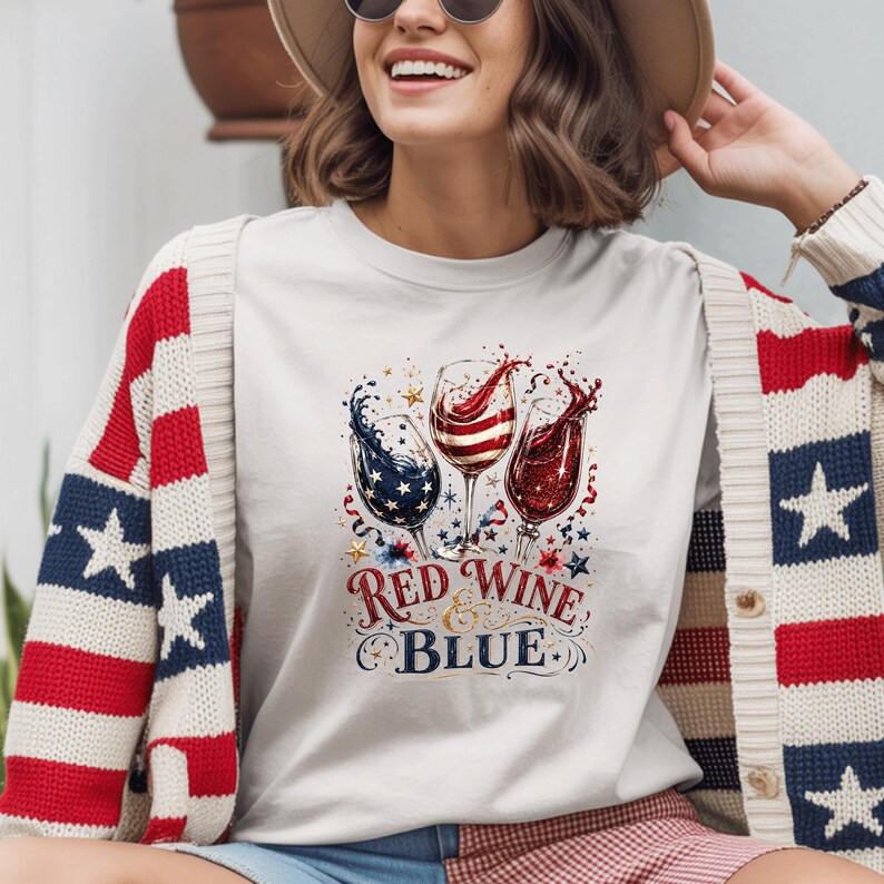 May include: A white t-shirt featuring a graphic of three wine glasses filled with red, white, and blue liquid, accented with stars and swirls. The text "RED WINE & BLUE" is printed below the glasses. The shirt is worn under a red, white, and blue cardigan.