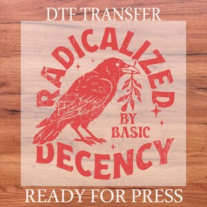 May include: Red DTF transfer with a distressed design featuring a raven holding a sprig. Text reads "RADICALIZED BY BASIC DECENCY" and "READY FOR PRESS". The design is on a transparent background.
