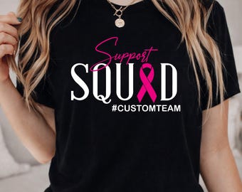 May include: A black t-shirt featuring the text Support SQUAD in white and pink, with a pink breast cancer awareness ribbon replacing the letter A. Below the main text, the hashtag #CUSTOMTEAM is printed in white capital letters.