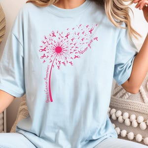 May include: A light blue t-shirt featuring a pink graphic of a dandelion where the seeds are small breast cancer awareness ribbons. The stem of the dandelion includes the text Breast Cancer Awareness and the phrase Never give up.