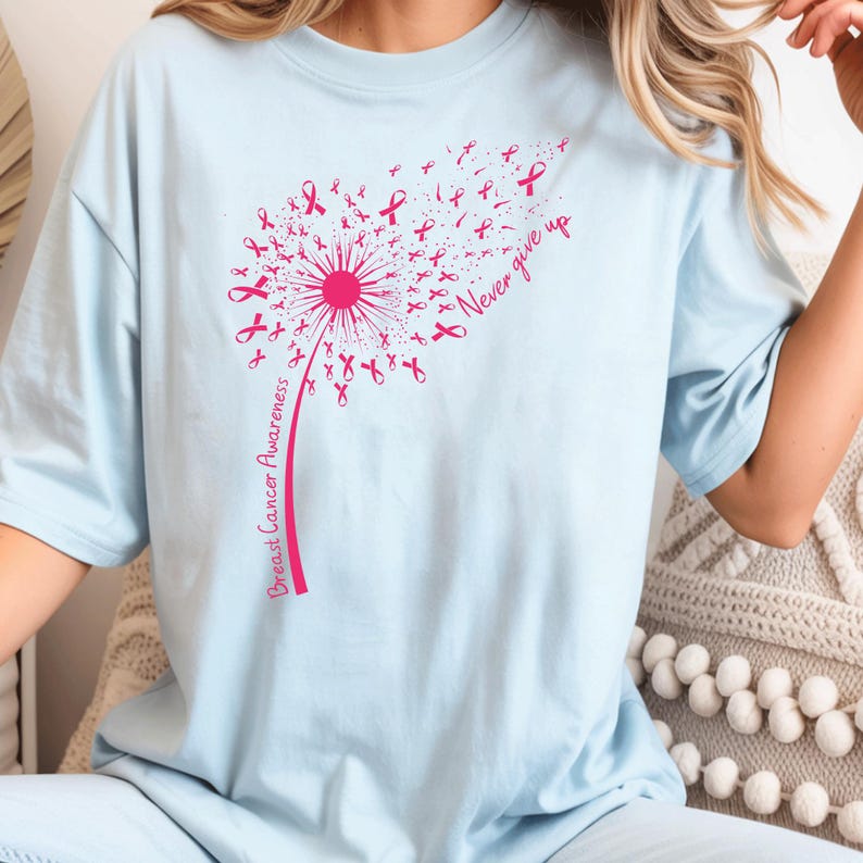 May include: A light blue t-shirt featuring a pink graphic of a dandelion where the seeds are small breast cancer awareness ribbons. The stem of the dandelion includes the text Breast Cancer Awareness and the phrase Never give up.