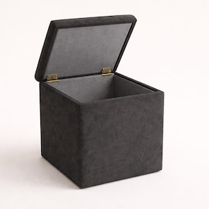 May include: A dark gray, cube-shaped storage ottoman with a hinged lid. The lid is open, revealing the interior. The ottoman has a soft, textured appearance and is set against a white background. The hinges are gold-colored.
