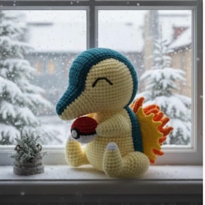 May include: A handmade Cyndaquil crochet plushie, featuring yellow, teal, and orange colours, clutching a red and white Poké Ball. The plush is positioned on a windowsill, with a snowy winter landscape visible through the window. A small potted plant is also present.