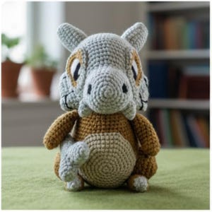 Puede incluir: Un peluche de Cubone hecho a mano con ganchillo en gris, marrón y beige. El peluche presenta una cabeza con forma de calavera, un cuerpo redondo y brazos y piernas pequeños. El juguete está sobre una superficie verde.