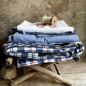 Könnte beinhalten: Ein Stapel gefalteter Stoffe in Blau- und Weißtönen, darunter Vichy- und Karomuster, liegt auf einem kleinen Holzschemel. Ein weißes Tuch mit roter Stickerei und eine Holzspule mit Faden liegen obenauf. Eine Kordelrolle liegt auf dem Boden.