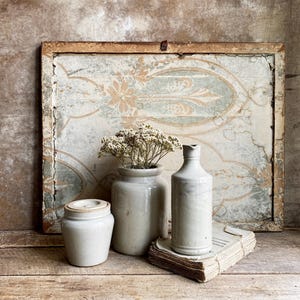Könnte beinhalten: Ein antikes Stillleben mit einem verwitterten Holzrahmen mit floraler Tapete und drei Keramikgefäßen. Ein kleines Glas mit Deckel, eine Vase mit Trockenblumen und eine hohe Flasche stehen auf einer Holzoberfläche.