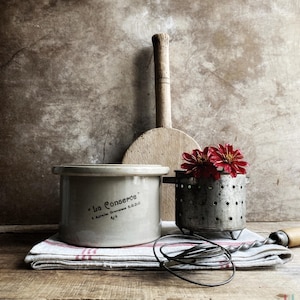 Peut inclure: Un pot en céramique beige avec l'inscription La Conserve et S.G.D.G. sur le côté, une spatule en bois, une passoire en métal avec des fleurs rouges et un fouet en métal posés sur un linge blanc à rayures rouges. Les objets sont regroupés.