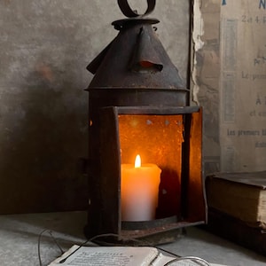 May include: A weathered, dark brown metal lantern with a conical top and a circular handle, containing a lit white pillar candle. In the foreground, a pair of round, thin-rimmed metal spectacles rests on an open antique book with printed text.