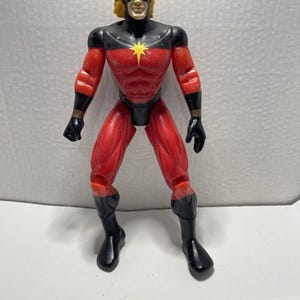 May include: A red and black action figure of a superhero with a yellow star on the chest. The figure has a black mask, yellow hair, and black gloves and boots. The figure is standing with its legs apart.