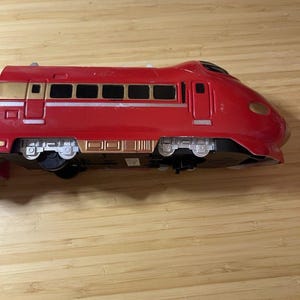 May include: A red toy train with gold accents. The train has multiple windows and detailed wheels. The front of the train is rounded, and the overall design suggests a high-speed train. The toy is made of plastic.