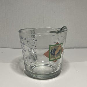 May include: A clear glass measuring cup with a handle and a spout. The cup has black measurement markings in milliliters and deciliters. A rooster graphic is on the front. The cup is designed for kitchen use.