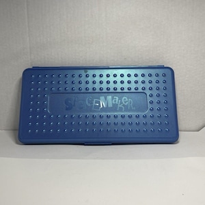 Puede incluir: Una caja de plástico azul con la palabra "SPACEMAKER" en relieve en la tapa. La caja tiene forma rectangular con esquinas redondeadas y está cubierta de pequeñas hendiduras circulares espaciadas uniformemente. La caja está cerrada.