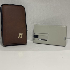 May include: A brown leather case with a black zipper and the word "Ranging" printed in gold. Next to it is a light gray rectangular device with the word "Feet" printed on it.
