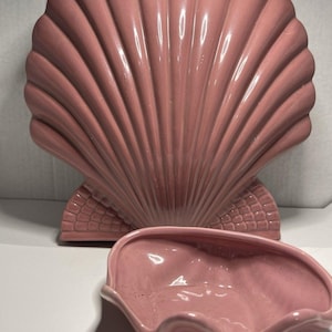 May include: A pink ceramic seashell-shaped decorative set. The set includes a large, upright shell and a smaller, matching dish. The shell has a ribbed design, and the dish has a wavy edge. The set is a soft, muted pink color.