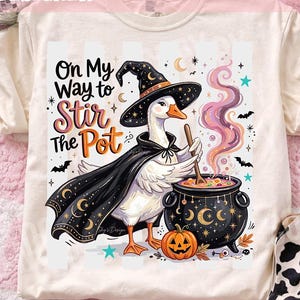May include: A cream-colored t-shirt featuring a graphic of a white goose wearing a black witch hat and cape, stirring a black cauldron with a wooden spoon. The text reads "On My Way to Stir the Pot" with pink and orange smoke, bats, and stars.