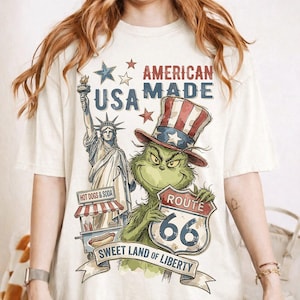 May include: Cream-colored t-shirt with a graphic featuring the Statue of Liberty, a green character in a patriotic hat, and Route 66 signage. Text includes "American Made," "USA," and "Sweet Land of Liberty."