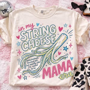 May include: A cream-coloured t-shirt featuring a playful graphic. The design includes the words "in my STRING CHEESE MAMA era" and a string cheese illustration. The text "overwhelmed, touched out, but exactly who I'm meant to be" is also present.