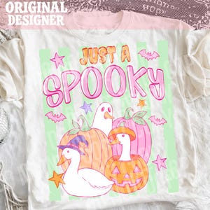 Puede incluir: Una camiseta blanca con un estampado gráfico que incluye el texto JUST A SPOOKY en naranja y rosa. El diseño muestra dos ocas blancas, una de ellas con un sombrero de bruja morado, junto a calabazas, un fantasma, murciélagos y estrellas.