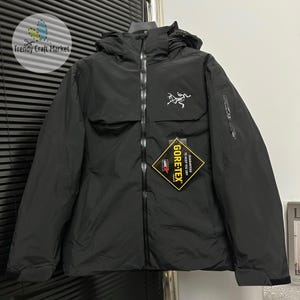 Può includere: Una giacca nera con cappuccio, dotata di cerniera frontale, logo bianco sul petto e tasca con zip sulla manica. Un'etichetta GORE-TEX gialla e nera a forma di rombo riporta il testo GUARANTEED TO KEEP YOU DRY.
