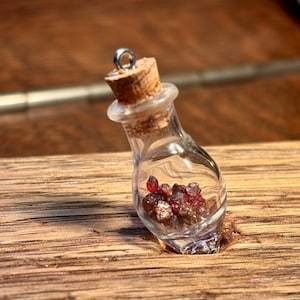 May include: A small, clear glass bottle with a cork stopper, filled with small, dark red gemstones. The bottle has a silver loop attached to the cork. The bottle is tilted slightly, and the gemstones are visible inside. The bottle is on a wooden surface.