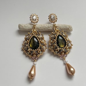 May include: A pair of elaborate gold earrings. Each earring has a teardrop-shaped, dark green gemstone, encircled by gold beads and small pearls. A small, clear gemstone is set at the top. The earrings dangle from a gold setting and have a pear-shaped, peach-coloured bead at the bottom.