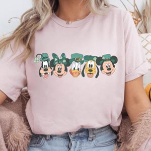 Puede incluir: Camiseta rosa claro con un diseño de cinco personajes de dibujos animados con sombreros verdes del Día de San Patricio. Los personajes son Goofy, Minnie Mouse, Donald Duck, Pluto y Mickey Mouse. El diseño está centrado.