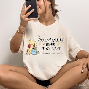 May include: Cream-colored t-shirt featuring a graphic of Winnie the Pooh holding a honey pot. The text "You can call me Hunny if you want" is printed in black. The design includes yellow stars and bees.