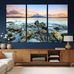 May include: A triptych canvas print depicting a coastal landscape at sunset. The artwork features a rocky shoreline with water, a mountain, and a colorful sky with clouds. The color palette includes blues, oranges, and grays.