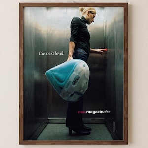 May include: A framed advertisement poster featuring a person in a black turtleneck and dark jeans standing in an elevator, holding a translucent blue computer monitor. The text reads "the next level." and "macmagazinabo" in a dark wooden frame.