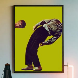 May include: A framed art print showing a person in a denim shirt and black trousers, bending backward against a bright yellow backdrop. The artwork is displayed above a white cabinet with a neon lamp and a vintage metronome.