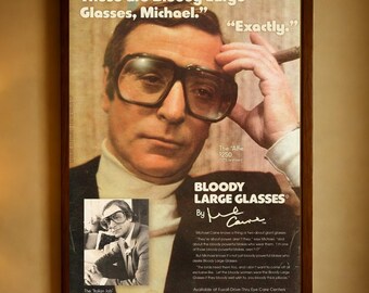Op de afbeelding: Een ingelijste advertentie voor "Bloody Large Glasses" met een man met een grote, donkeromrandde bril. De advertentie bevat de tekst "Those are Bloody Large Glasses, Michael." en "Exactly."
