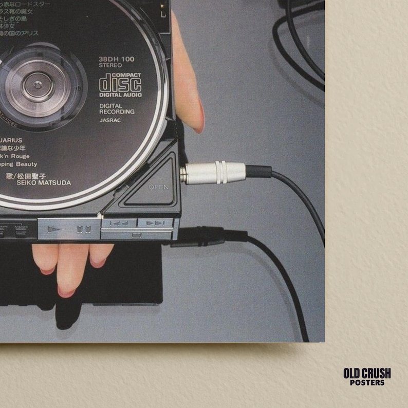 Op de afbeelding: Een zwarte cd-speler met een zilveren stekker verbonden met een zwarte kabel. De cd-speler is open en onthult een compact disc met de tekst "COMPACT disc DIGITAL AUDIO". De afbeelding heeft Japanse tekst.