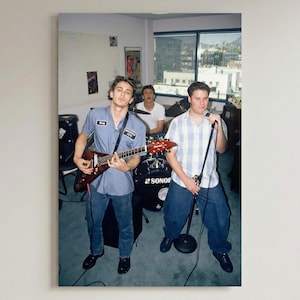 May include: A framed print showing a band playing music in a room. The guitarist is wearing a blue work shirt and jeans, the singer is holding a microphone, and the drummer is in the background. The singer is wearing a plaid shirt and jeans.