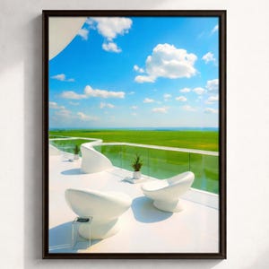 May include: Framed art print featuring a modern outdoor scene. The image showcases a white patio with two curved chairs, a small table, and potted plants. A glass railing overlooks a vibrant green field under a bright blue sky with fluffy white clouds.