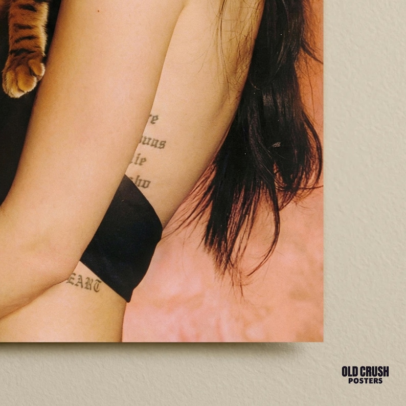 May include: Close-up of a person's arm with tattoos. One tattoo reads "HEART" and another features text in a gothic font. A black garment is visible. A portion of a cat's paw is in the upper left corner. The background is a light pink.