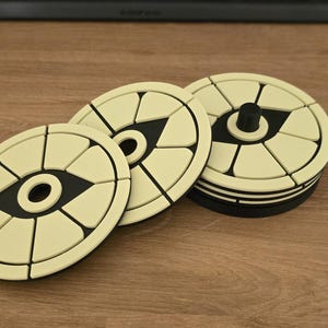 May include: A set of three circular coasters featuring a cream-coloured top surface with a black geometric eye-shaped design in the centre. The coasters have a segmented pattern and a solid black base, with one coaster featuring a small black central knob.