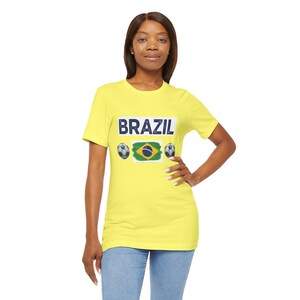 Puede incluir: Una camiseta amarilla con la palabra "BRAZIL" en azul. Debajo, la bandera brasileña y dos balones de fútbol. La camiseta la lleva una persona.
