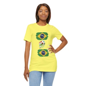 Puede incluir: Una camiseta amarilla con tres diseños: dos banderas brasileñas y un balón de fútbol. Las banderas son verdes con un rombo amarillo y un círculo azul con estrellas. El balón es blanco con rayas coloridas.