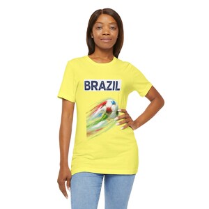 Puede incluir: Una camiseta amarilla con la palabra "BRAZIL" en letras azules. Debajo del texto hay un gráfico de un balón de fútbol con rayas rojas, verdes y azules. La camiseta la lleva una persona.