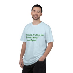 Könnte beinhalten: Hellblaues T-Shirt mit dem Zitat "Im Krieg ist die Wahrheit das erste Opfer." von Aischylos in grüner Schrift. Das Shirt hat einen Rundhalsausschnitt und kurze Ärmel.